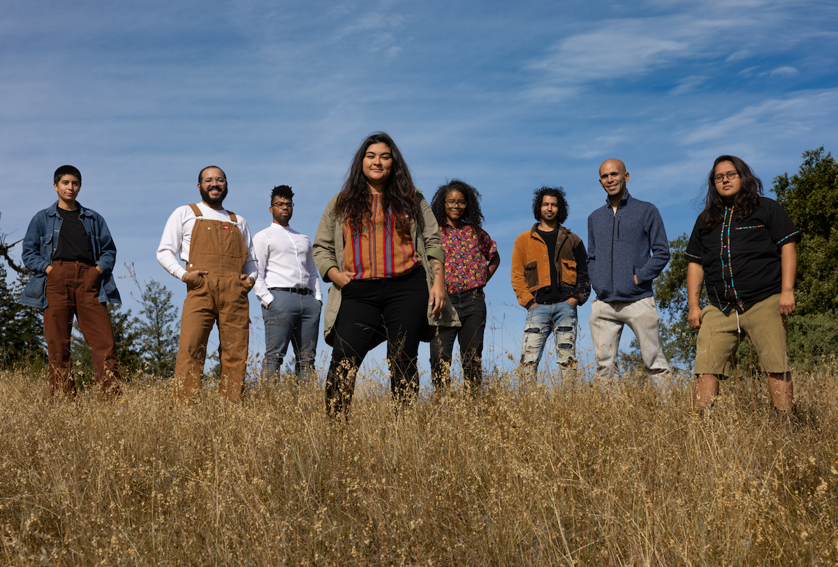 eight members of Shelterwood Collective, a Black, Indigenous, and LGBTQ-led community forest and retreat center. they stand in a row, looking directly at the camera, against a bright blue sky and dry yellow grass. their facial expressions show happiness or contentment.