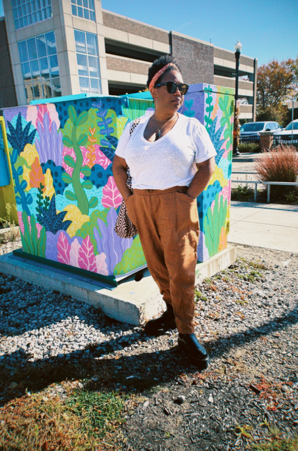 Standing in a parking lot in front of a brightly painted structure, Carmen wears brown pants, a white t-shirt, and her hair is pinned up with a coral-colored headband.