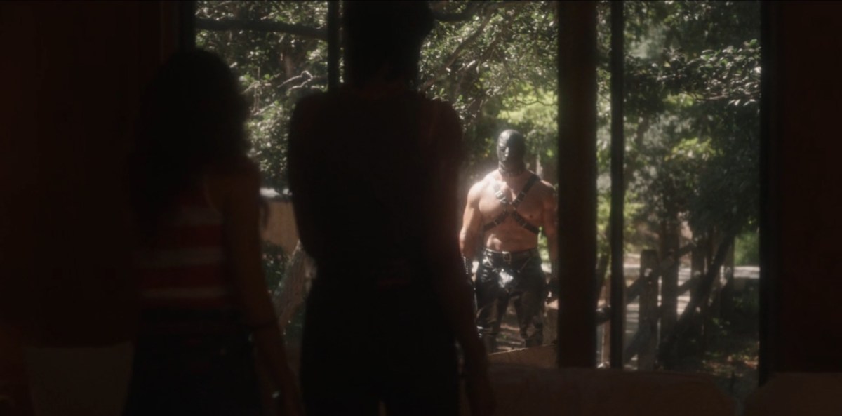 Two women backlit look out their big window at a buff man in a leather mask.