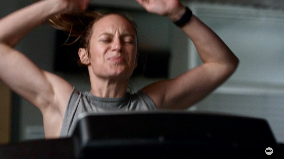 Maya is sweaty, running on a treadmill, her hands above her head.