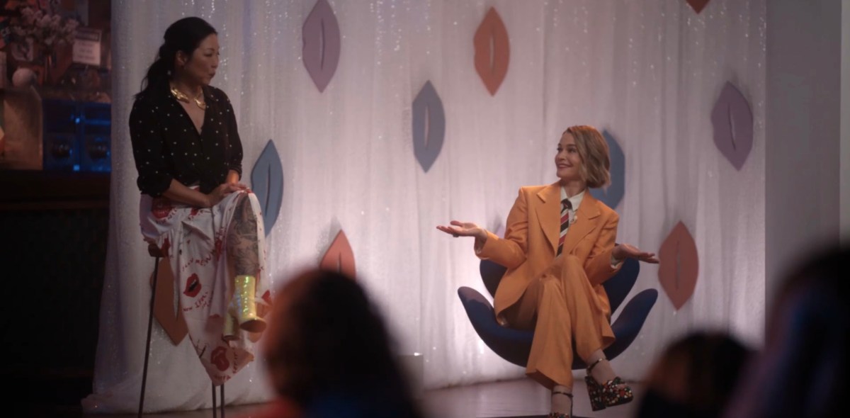 Alice in her orange suit in a funny chair doing a shrug and Margaret Cho smiling at her from a stool