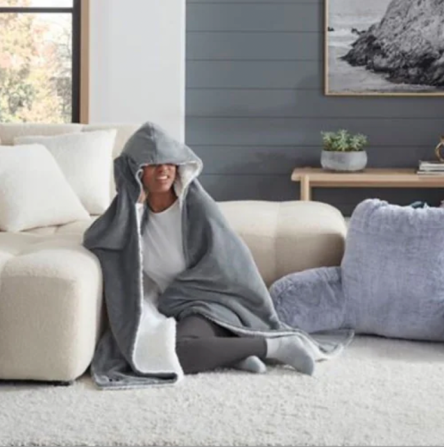 a black woman in a gray hooded throw sitting on the floor against a cream couch. there is a gray pillow next to her on the floor and a gray wall behind her