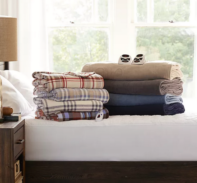 a pile of plaid print throws on a white bed with a brown lamp on a brown side table