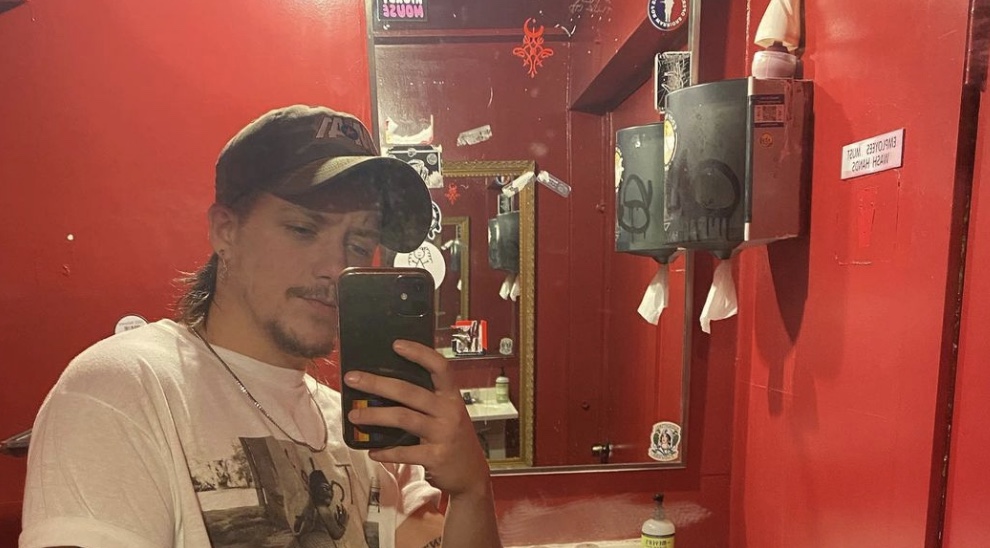 Daniel Davis Aston, a man in a baseball cap and white t-shirt with a chain, takes a bathroom selfie with his phone