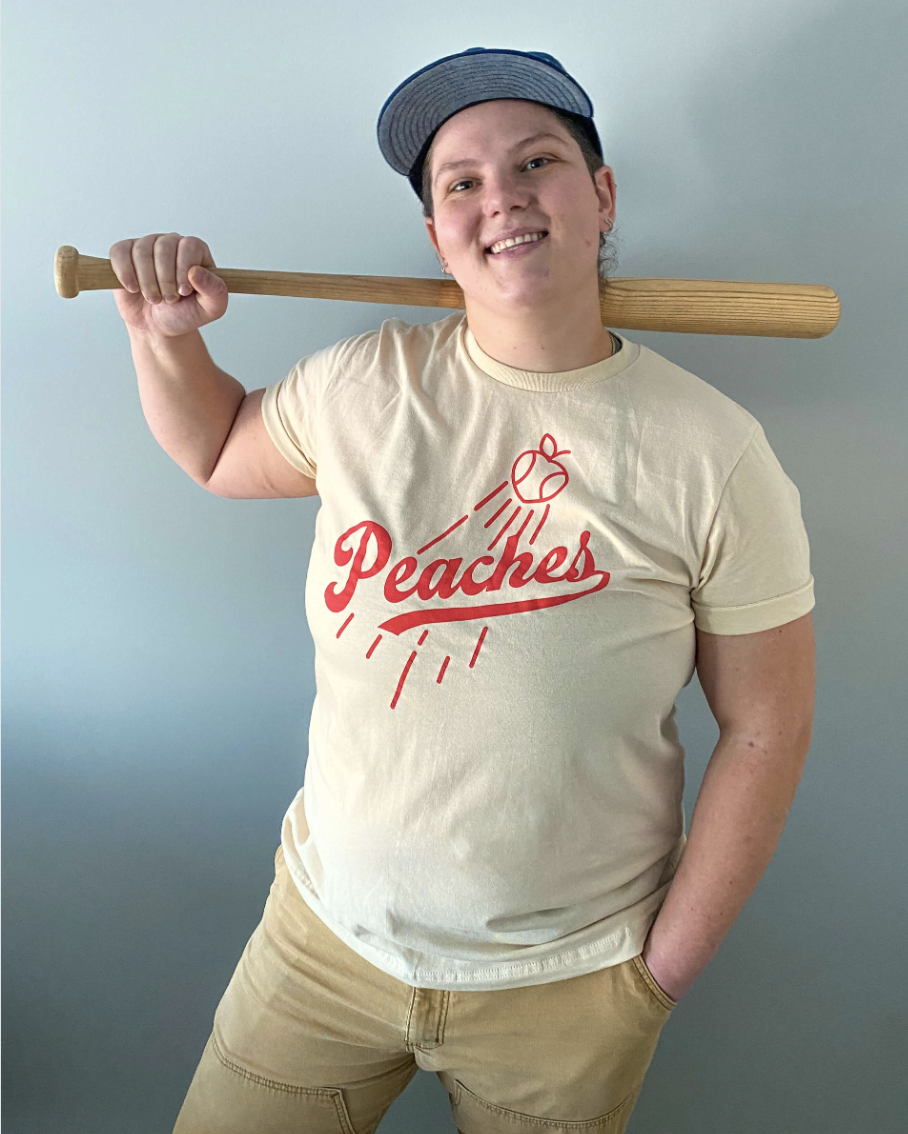 Model Jenny is 5'9" and a 38DDD, wearing size XL. The Rockford Peaches Tee is a retro print of the word "Peaches" in a baseball typeface. There is also an outline of a peach with lines on it to resemble a baseball being thrown up into the air. The print is on a soft cream color shirt that is unisex sizing and super soft. The Rockford Peaches is one of the main baseball teams in the 2022 tv show "A League Of Their Own." Wear this to all your gaymes and root for the team that plays for your team!