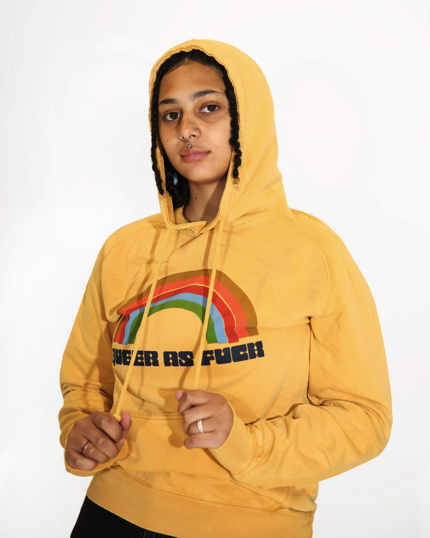 The model Grey is wearing the mustard yellow Queer As Fuck Vintage Hoodie in size S. The hoodie has a graphic of a rainbow in retro colors and the words "QUEER AS FUCK" underneath the rainbow.