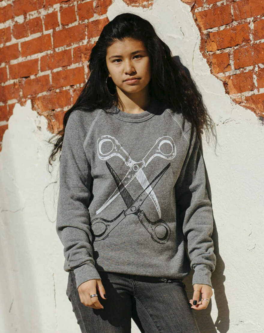 The model Jordan is wearing the grey Basic Scissoring Sweatshirt in size S. The sweatshirt is grey and has a graphic of two scissors, blades open and intertwined. The top scissor is white and the bottom is black.
