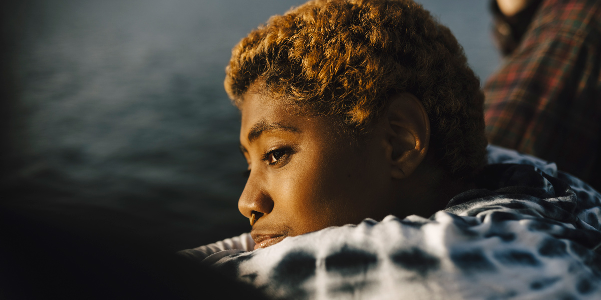 A Black nonbinary trans person with a short natural hairstyle, died blonde, and a septum piercing, is bundled underneath a black and white blanket, and is looking out over a dark blue ocean. Their face seems to be contemplative.