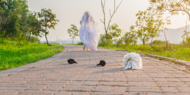 A bride in a long wedding gown and veil is running away down a brick path. Trees are visible on either side of her, and hills are in the distance. Her black high heels and white bouquet are on the ground.