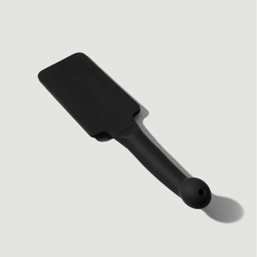 A black, silicone paddle, which has a long handle with a bulbous end with a small hole in it is against a light grey background. The paddle casts a small shadow under the handle.