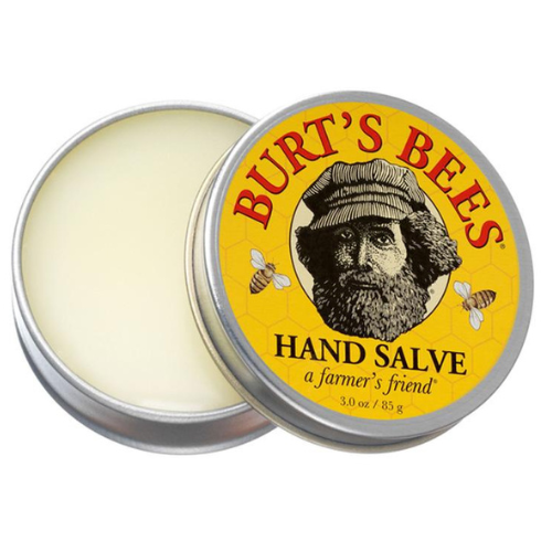 An open, circular metal tin containing a white hand salve is against a white background. The top of the tin is yellow. Red text reads, "Burt's Bees" and black text reads, "Hand Salve - a farmer's friend." There is a black and white drawing of a bearded man in a bucket hat in the center of the tin. Drawings of bees are on either side of the man's face.