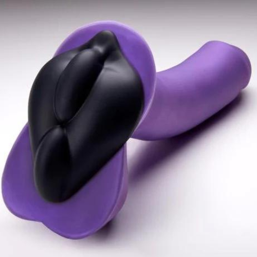 A purple dildo with a wide base with balls faces away from the camera against a grey background. A black cushion that's shaped like a vulva with ridges down the center sticks to the base of the dildo.