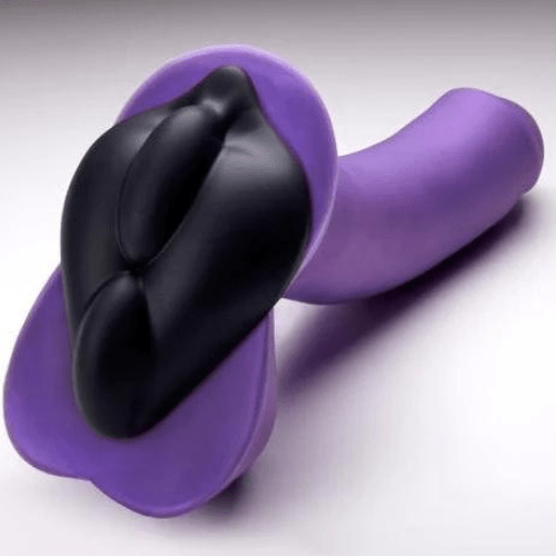 A purple dildo with a wide base with balls faces away from the camera against a grey background. A black cushion that's shaped like a vulva with ridges down the center sticks to the base of the dildo.