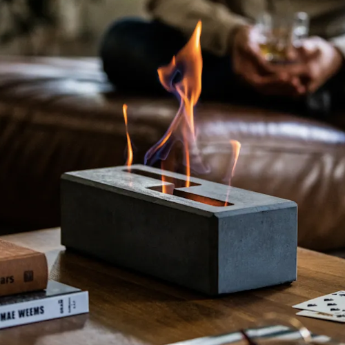 A concrete block with a narrow opening emits flames. The block sits on a coffee table with books and cards. Behind the coffee table, part of a brown leather couch is visible. A person in jeans and a sweater sits on the couch holding a glass.