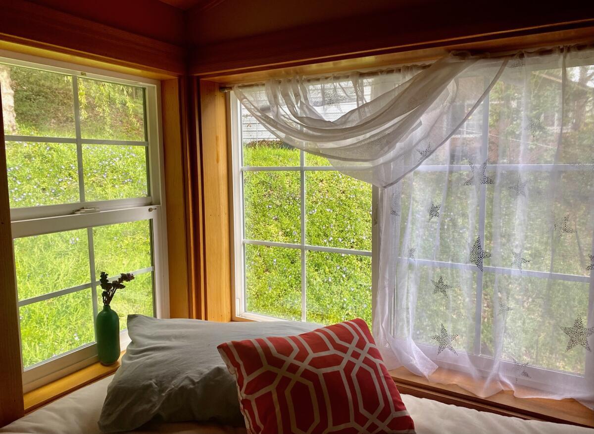 Loft at Mugworts: Large windows looking out onto green trees, transparent curtains tucked to the side, pillows on a bed right by the windoiws