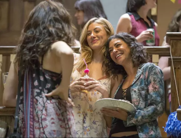 A still shot from Season 8, Episode 8 of Shameless shows Nessa and her girlfriend Mel standing in a crowded room. Nessa has chin-length way brown hair. She wears a multi-colored, snakeskin-patterned hoodie over a black tank top and rests her head on Mel's shoulder while holding a pie. Mel has long blonde hair and wear a white maxi dress with a floral pattern. She holds a beverage with a pink straw. Fiona stand with her back to the camera speaking to Nessa and Mel. Fiona has long, wavy brown hair and wears a dark blue, red, and white maxi dress with a floral pattern. She puts her pant on her hip.