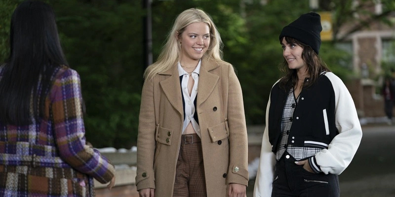 Kate and Lucy hold hands and lean against each other on the beach. They're sitting on a striped blanket. Kate is wearing khaki pants, a blue t-shirt with queen anne neckline. Kate has on no shoes. Lucy is wearing a light pink blouse with dark jeans and black boots.