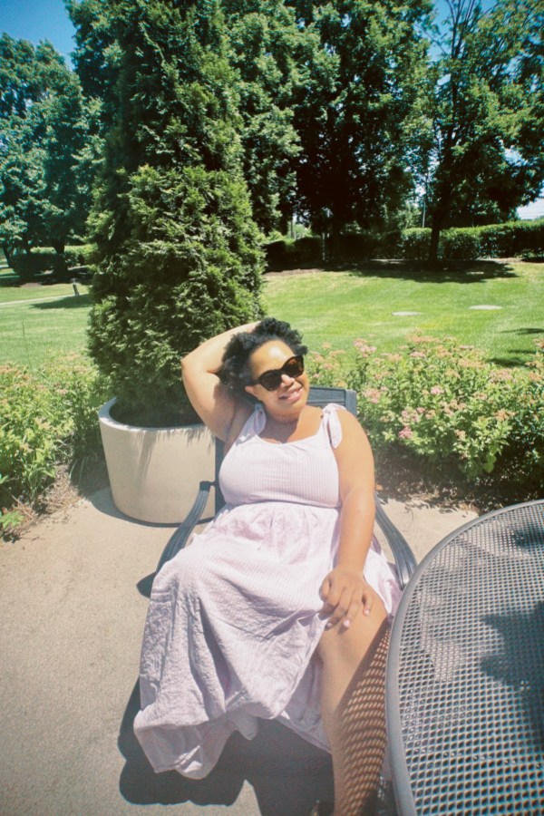 Carmen, a Black woman with glasses and natural hair, is sitting in a chair in a park in a light purple dress and sunglasses