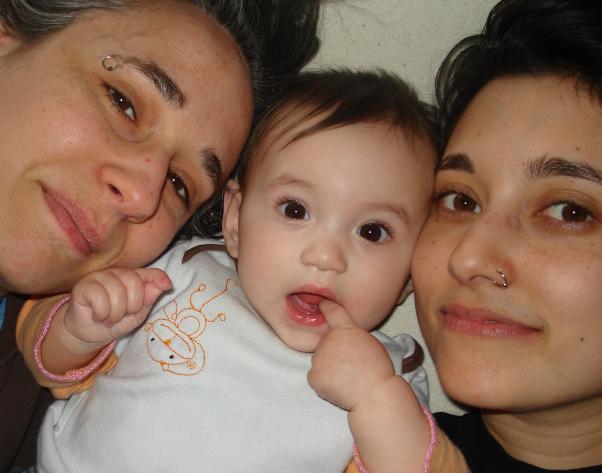 Tracy and Mia lie with their very young baby between them. It's a close-up of their three faces. Tracy and Mia are smiling lightly and the baby has his finger in his mouth.