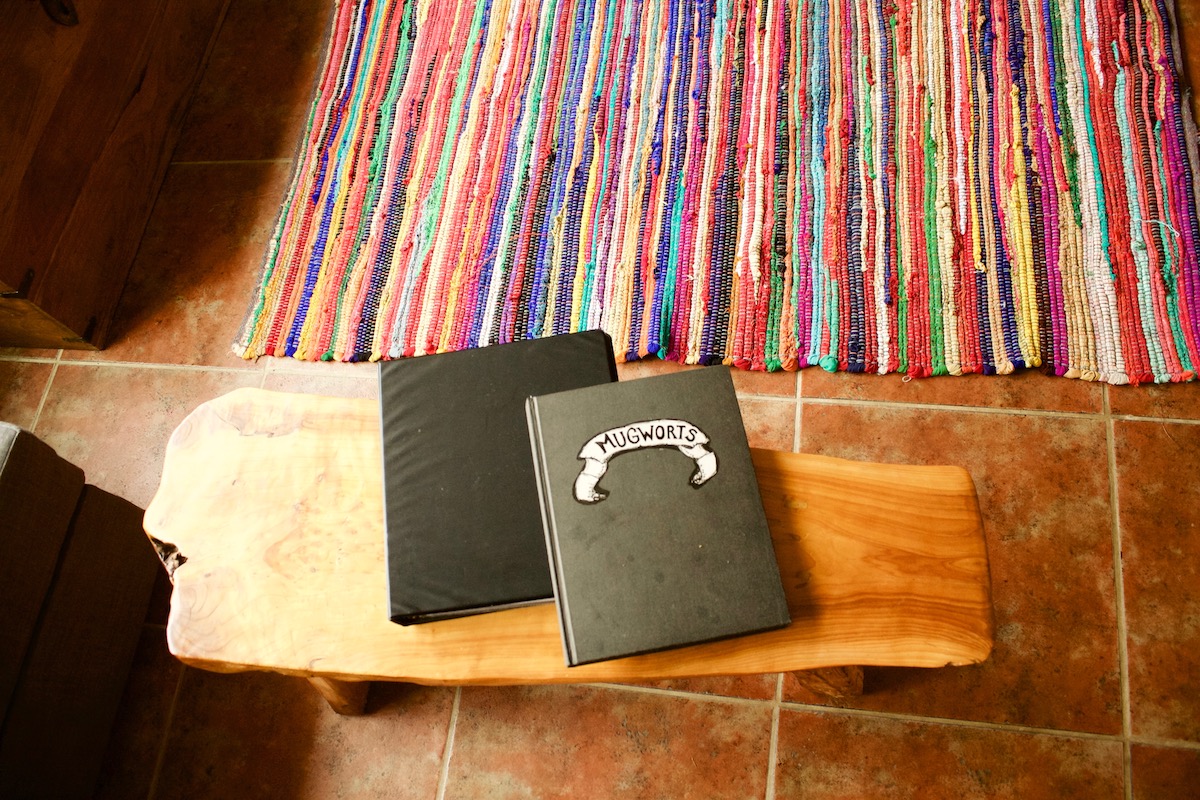 Black notebooks that say "Mugworts" on the front, on a wood bench next to a colorful rug