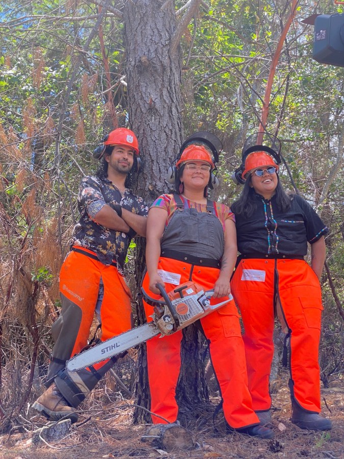 three members of Shelterwood Collective, a Black, Indigenous, and LGBTQ-led community forest and retreat center, in orange helmets and orange pants. one member holds a chainsaw; they all wear noise canceling headphones and they are all smiling.