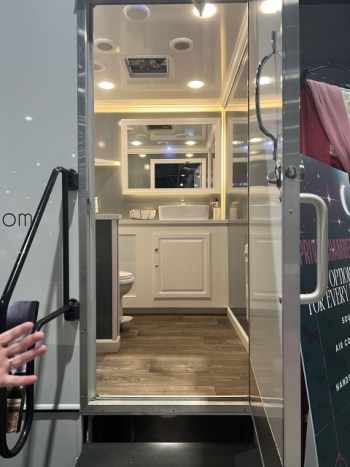 A white person's hand with an engagement ring gestures from the left of the image at the open door of a wedding trailer, which has wood flooring, a toilet, a white cabinet, a white sink, a mirror, bright overhead lighting, soap, and paper towels inside. The photo was taken at a Bridal Expo.