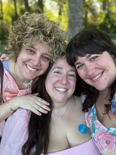 Vanessa, a white person with long, brown hair wearing pink clothing and a silver necklace, stands between two friends at a Bridal Expo. The friend on the left is a white person with curly blonde and grey hair who's wearing pink clothing and a silver bracelet. They put their hand on Vanessa's shoulder. The friend on the right is a white person with dark brown, shoulder-length hair and bands. They're wearing pink and green floral print clothing. All three smile.