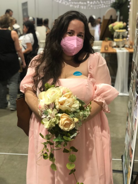 Vanessa, a white person with long, brown hair, stands in a crowded room at a Bridal Expo. She's wearing a pink dress with ruffles on the sleeves, a matching pink KN95 mask, and a blue sticker on her chest. She's holding a bouquet of white and pink roses.