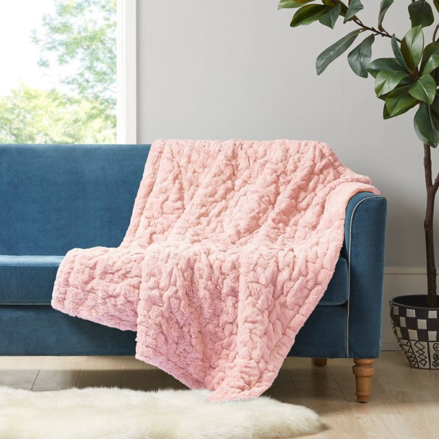 a soft pink blanket draped over a blue couch against a gray wall. there is an open window with trees