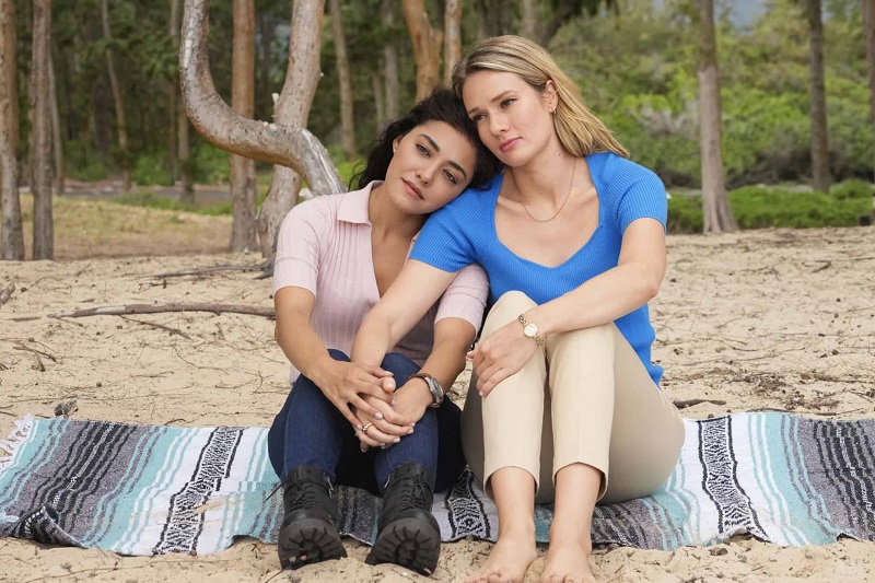 Kate and Lucy hold hands and lean against each other on the beach. They're sitting on a striped blanket. Kate is wearing khaki pants, a blue t-shirt with queen anne neckline. Kate has on no shoes. Lucy is wearing a light pink blouse with dark jeans and black boots.