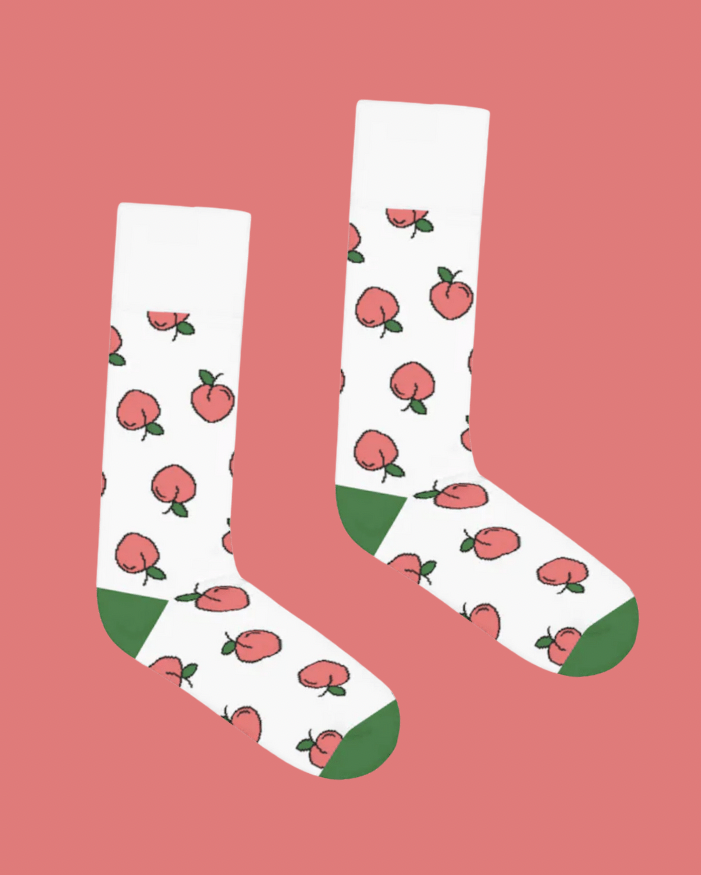 The Peaches Sock on a pink background. The sock is white and has a repeating print of a pink peach graphic. The peach is pink with a little green leaf, and all thinly outlined in black with a line for shine. The style is white cotton crew with a sporty green heel and toe color.