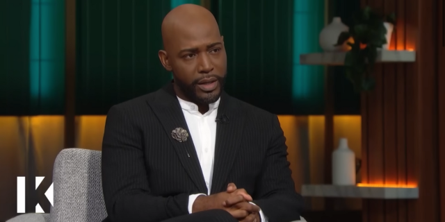 Karamo Brown sits on a chair on stage for his show, wearing a white button down and black suit.