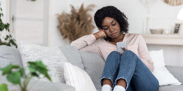 lonely Black woman looks at her phone with a thoughtful expression while sitting alone at home