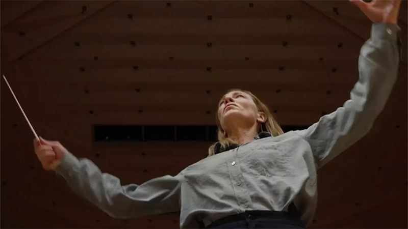 Cate Blanchett as Lydia Tar, with her arms spread wide, in a pristine blue button-down shirt