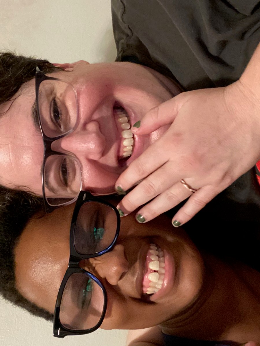 Sai, a black woman with short hair is wearing black glasses and smiling. Beth, a white woman with short hair is wearing glasses and smiling. She is holding up her left hand, a small rose gold band is on her ring finger