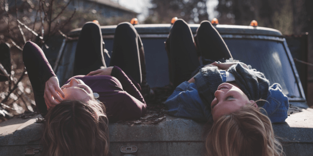 two young women lying in a truck bed looking at one another, wearing dark pants, scarves, and long sleeves