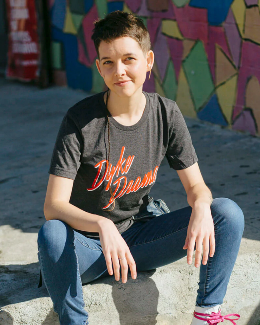 Model Janell is wearing the Dyke Drama Tee in size XS. She is 5'7" and her bra size is 34C. The tee is charcoal grey with the graphic "Dyke Drama" in a jagged red typeface with a white drop shadow.