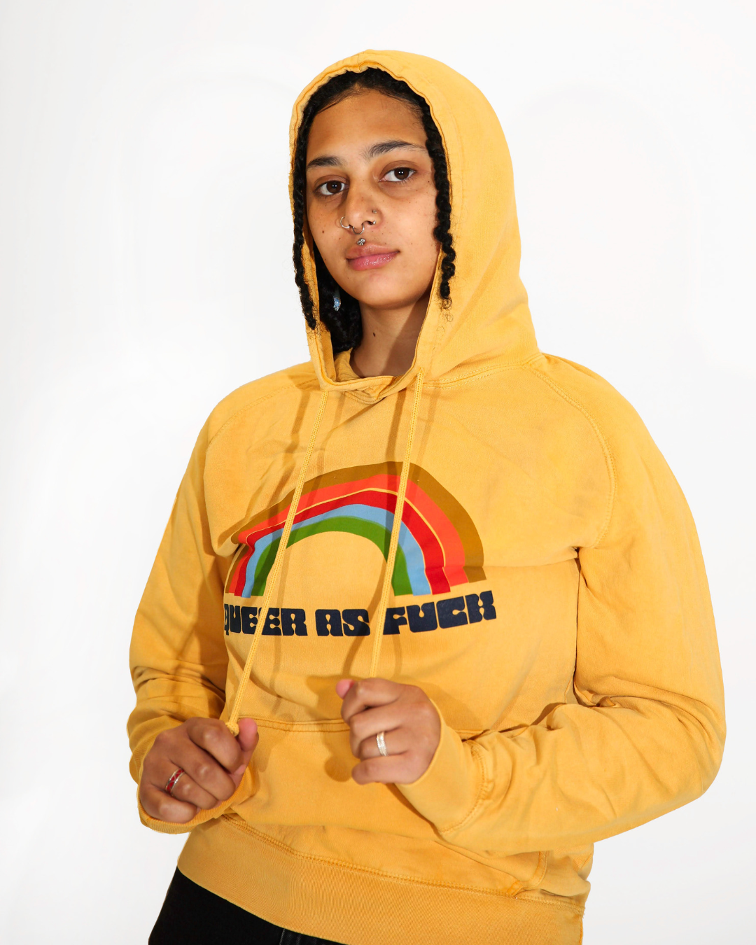Model Grey is wearing the Queer As Fuck Vintage Hoodie in size S. They are 5'4" and their bra size is 32D. The hoodie is mustard yellow with a four-band rainbow of retro 70s colors. Across the bottom of the rainbow is the text "Queer As Fuck" in a groovy funk typeface.