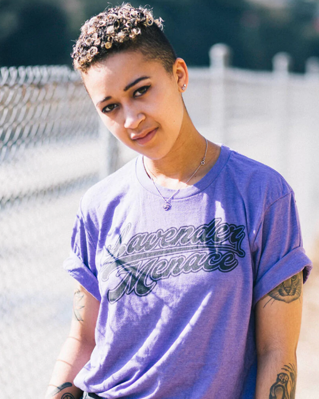 Model Savannah is wearing the Lavender Menace Tee in size L. They are 5'4" and their bra size is 34C. The tee is a faded purple with a dark grey graphic that says "Lavender Menace" in a retro sporty cursive typeface.