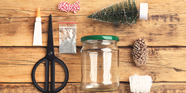 an image depicting the supplies needed for making snow globes at home. these include a glass jar, scissors, string, a plastic evergreen tree, glue, glitter and a pine cone