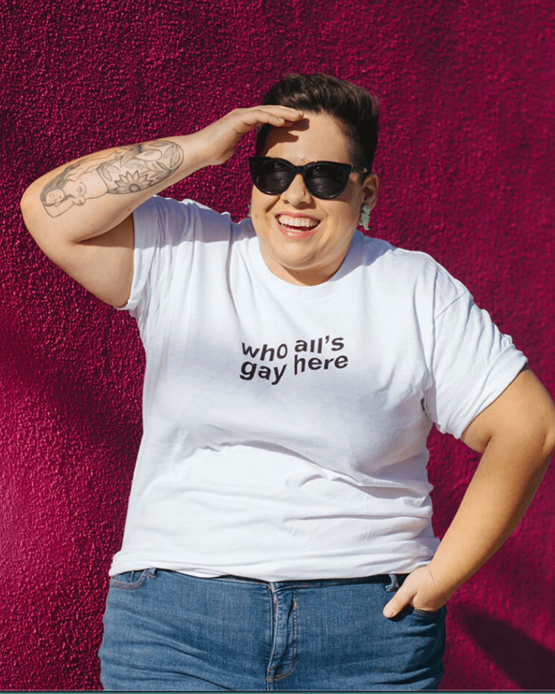 Model Dafne is wearing the Who All’s Gay Here White Tee in size XL. She is 5'1" and her bra size is 44D. The tee is white with black sans-serif text that says "Who All's Gay Here." The text is askew at a diagonal in two lines.  The design is by Erin Sullivan.