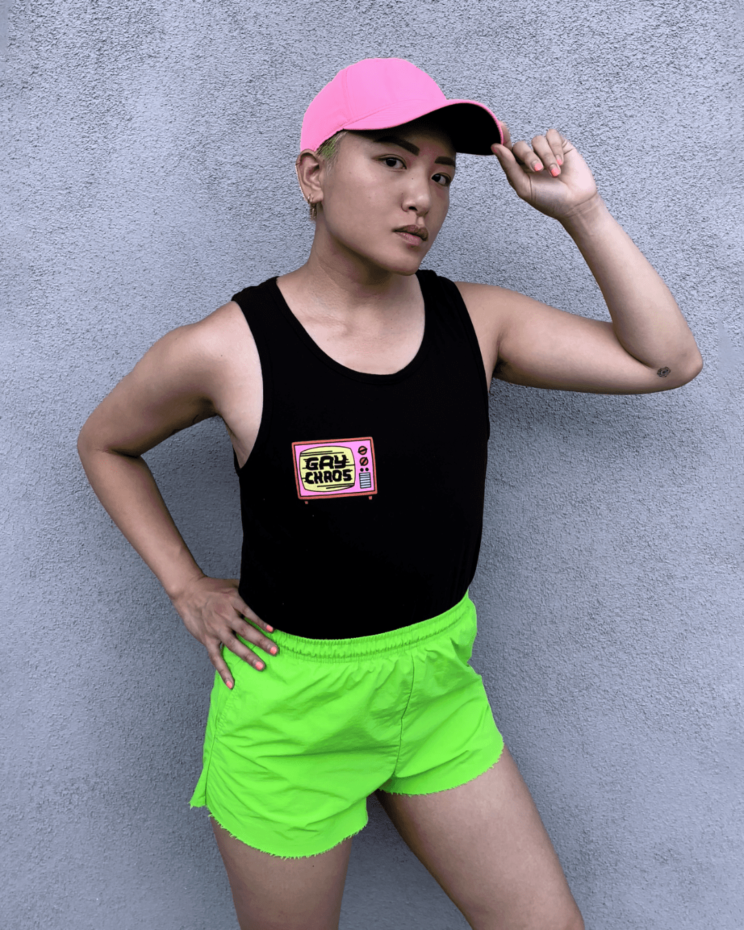 Model Viv is wearing the Gay Chaos Tank in size M. They are 5'6" and flat-chested. The tank is black with a unisex cut. On the right pocket is a pink and yellow graphic of a broken tv. On the tv screen is the text "Gay Chaos" in all caps. The graphic illustration was made by Betty Turbo.