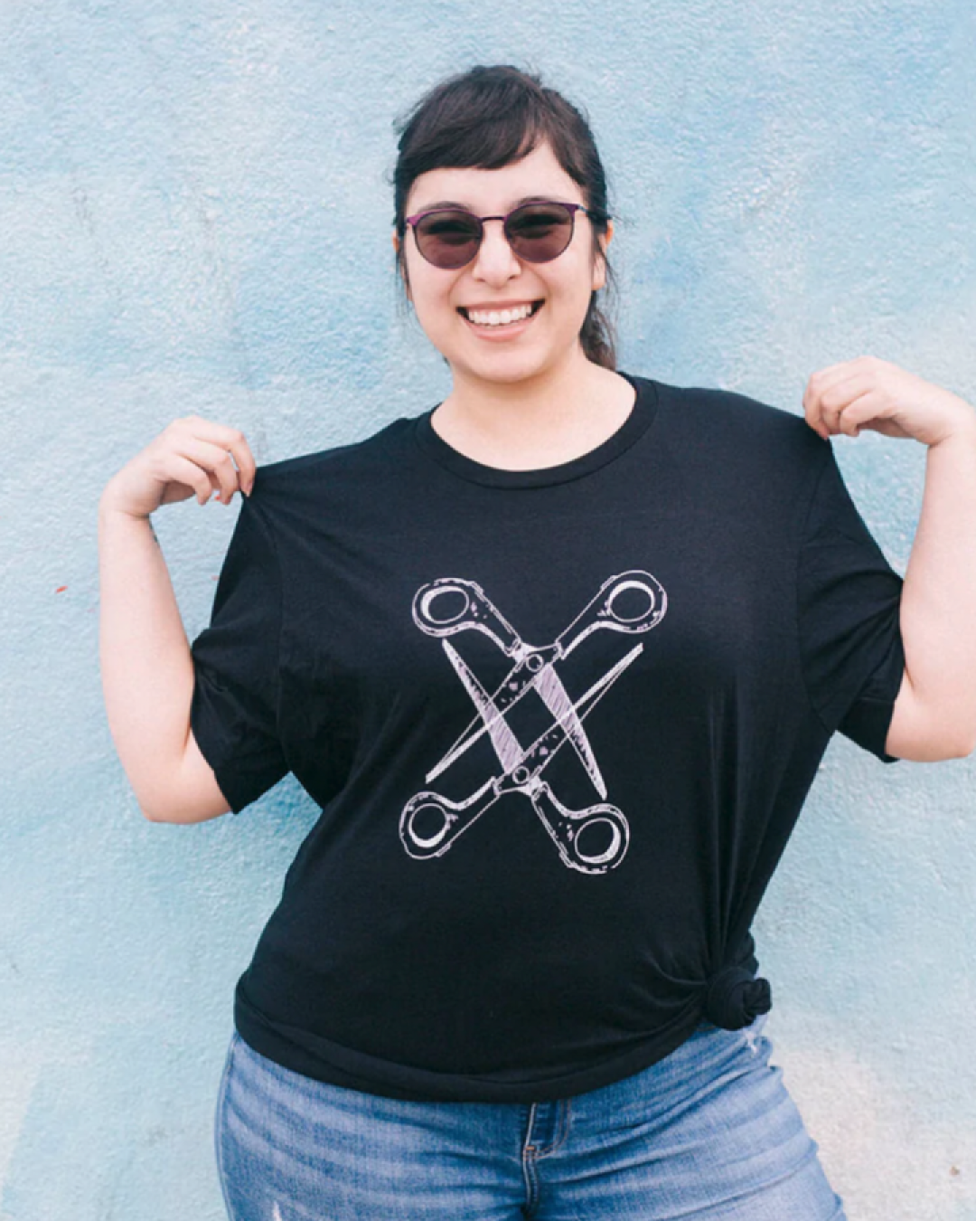 Model Julissa is wearing the Basic Scissoring Black Tee in size XL. They are 5'5" and their bra size is 36C. The tee is black with a graphic of two scissors in a vertical alignment. Their blades are open and overlapping each other. The graphic is outlined in white.