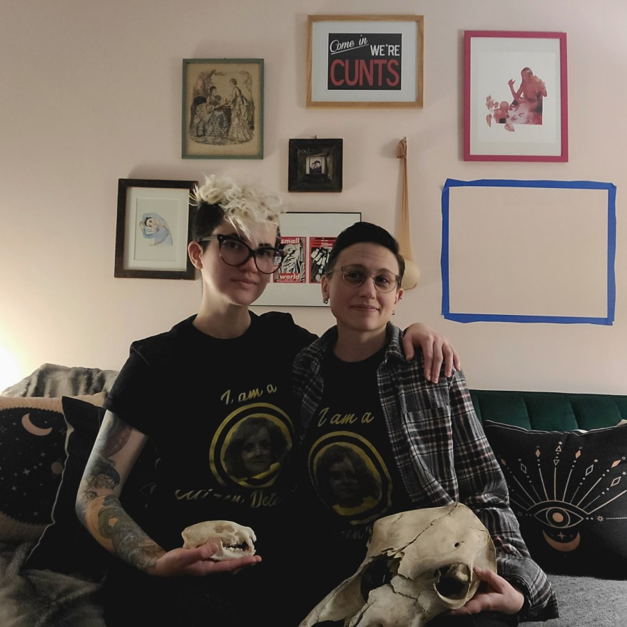Nico and Sadie each where "I'm a Citizen Detective" shirts and hold animal skulls, seated on their green velvet couch in front of their art collection. Nico's hair is bleached and shaved on the sides. Sadie's hair is short. They both wear glasses.