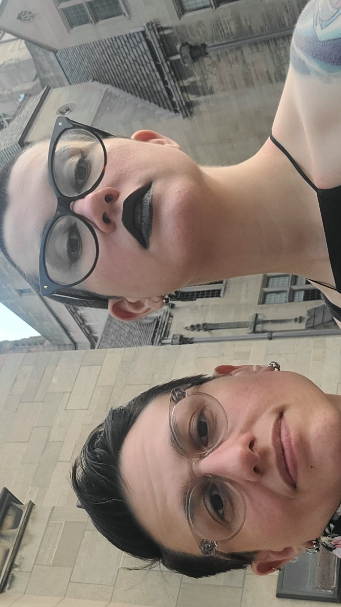 nico and sadie are dressed up and in front of a church. nico has a shaved head and is wearing a black dress and black lipstick. sadie is wearing a floral print button down shirt. they both wear glasses