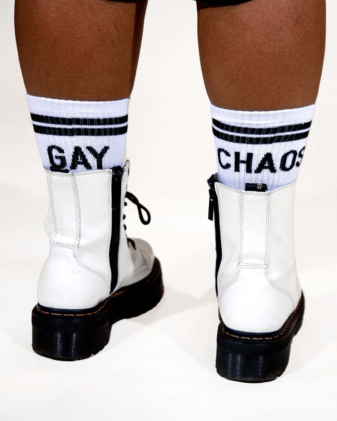 The GAY CHAOS Socks are white with black bands at the cuff. The socks have black text that say "GAY" on the back left cuff and "CHAOS" on the back right cuff. The style is ribbed cotton crew with compression and cushion. There is also black heel and toe color.