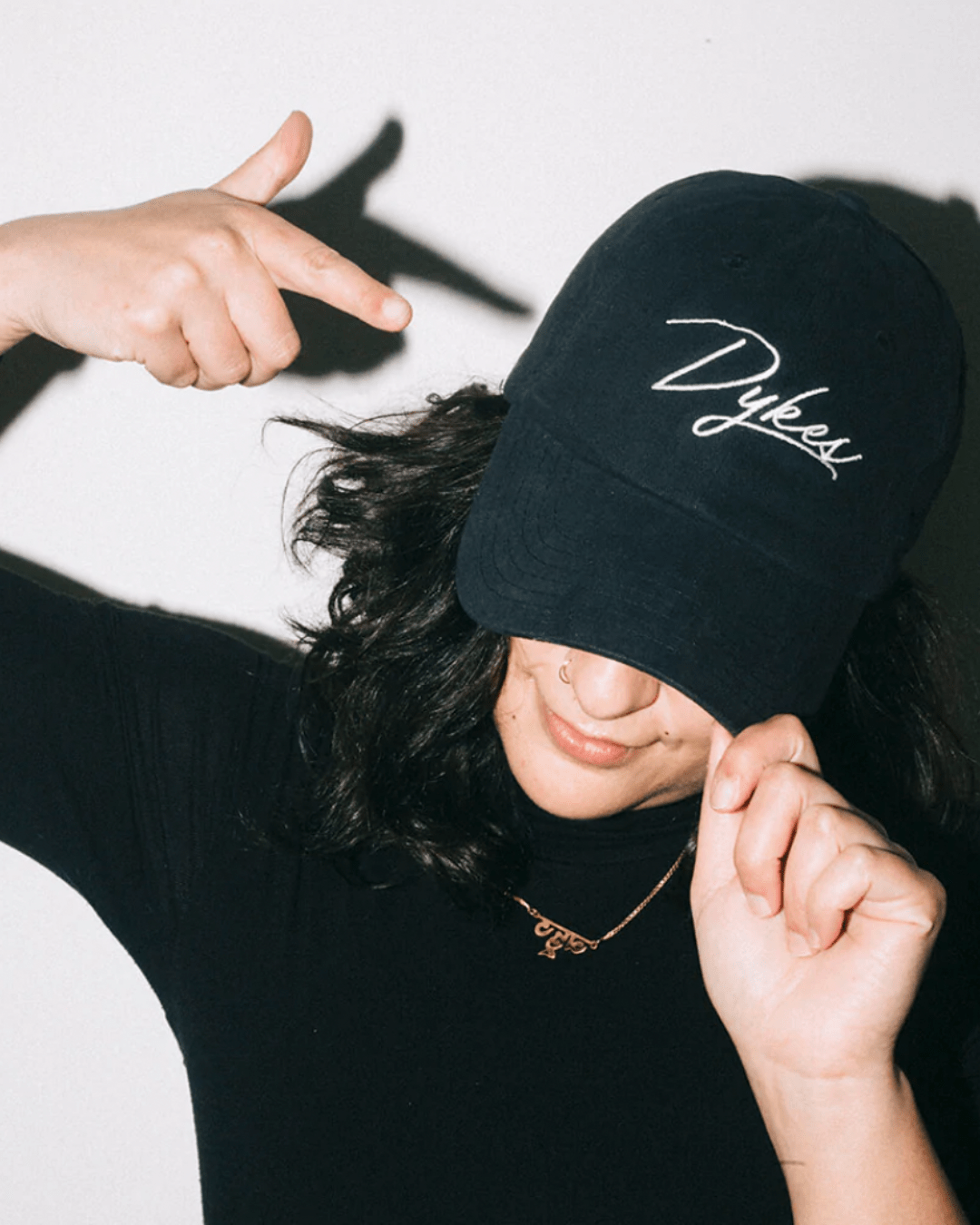 Model Sarah is wearing the Dykes Hat. The hat is a baseball cap style in black color with the text "Dykes" in white color to resemble a hand-drawn signature.