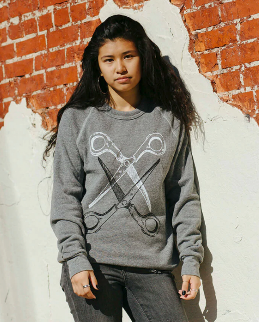 Model Jordan is wearing the Basic Scissoring Sweatshirt in size S. She is 5'3" and her bra size is 34B. The sweatshirt is grey with a graphic of two scissors in a vertical alignment. Their blades are open and overlapping each other. The top scissor is white and the bottom is black.
