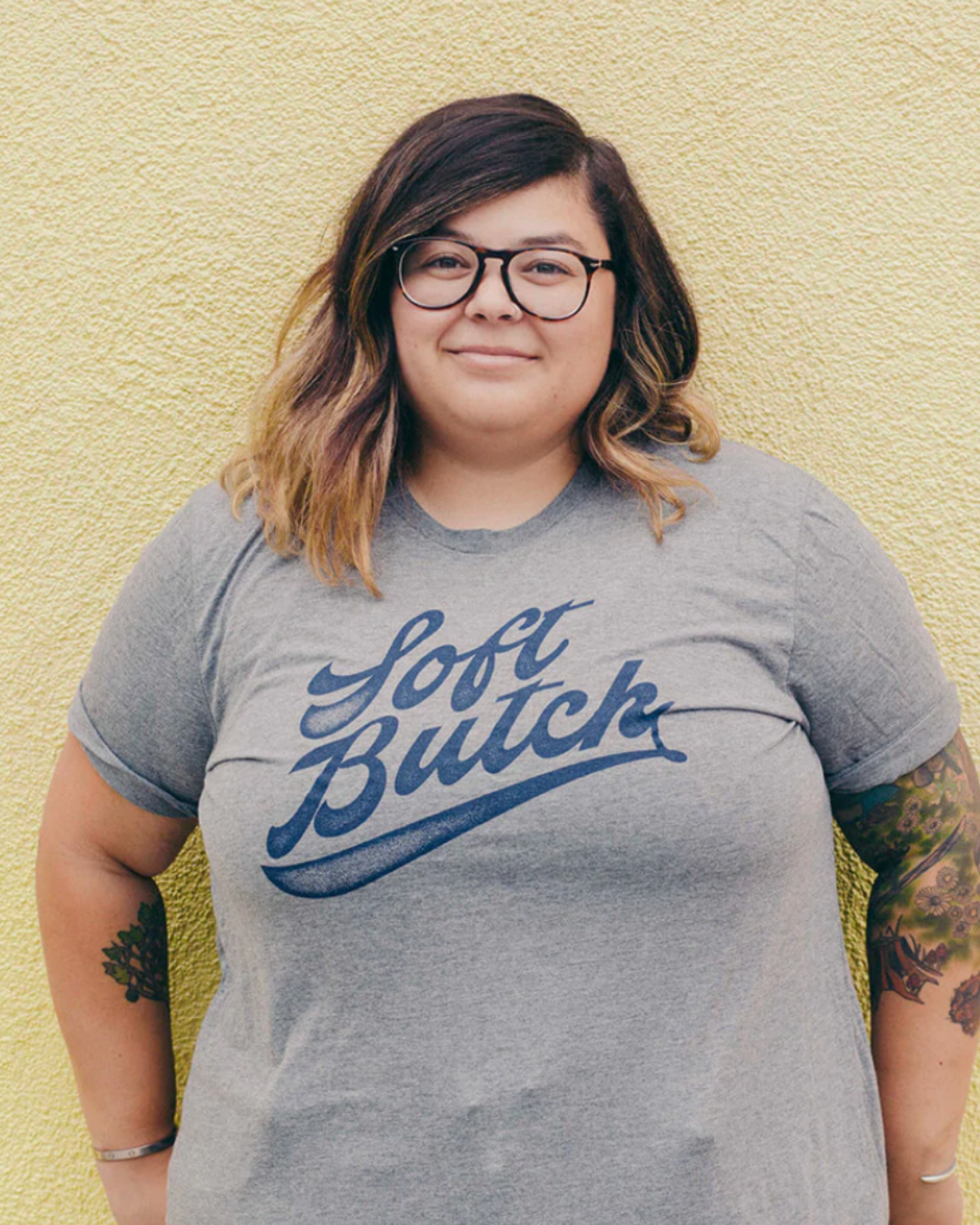 Model Chloe is wearing the Soft Butch Tee in size 2XL. She is 5'2" and her bra size is 38DD. The tee is grey a graphic type that says "Soft Butch" in navy blue retro sporty cursive.