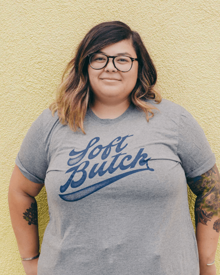 Model Chloe is wearing the Soft Butch Tee in size 2XL. She is 5'2" and her bra size is 38DD. The tee is grey a graphic type that says "Soft Butch" in navy blue retro sporty cursive.